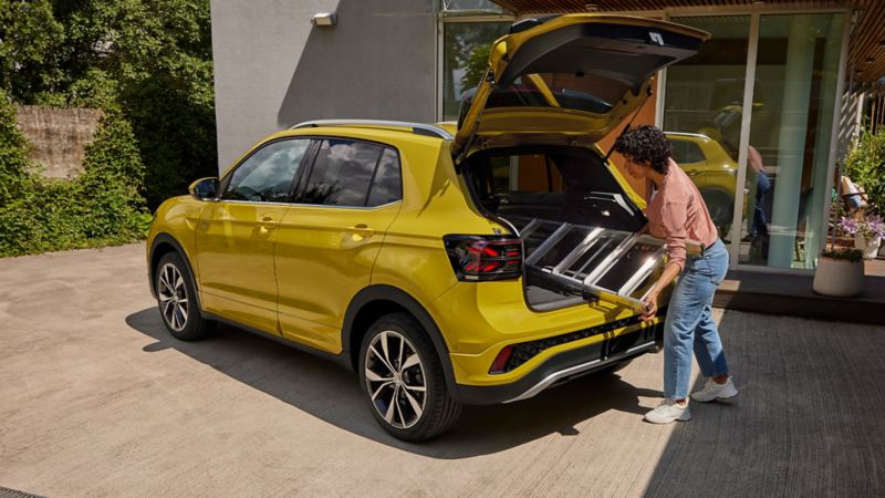 A young woman pushes a ladder through the open boot lid of a yellow VW T-Cross.