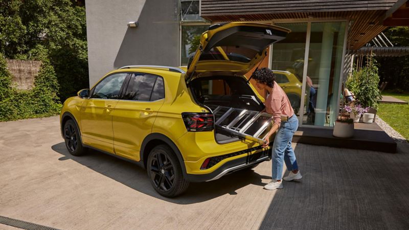 A young woman pushes a ladder through the open boot lid of a yellow VW T-Cross.