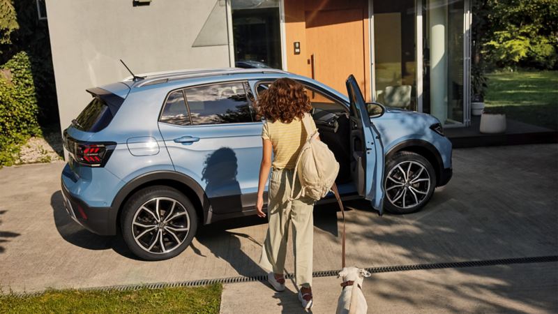 Une fille avec un chien se dirige vers un VW T-Cross bleu dans une allée. La porte passager est ouverte.