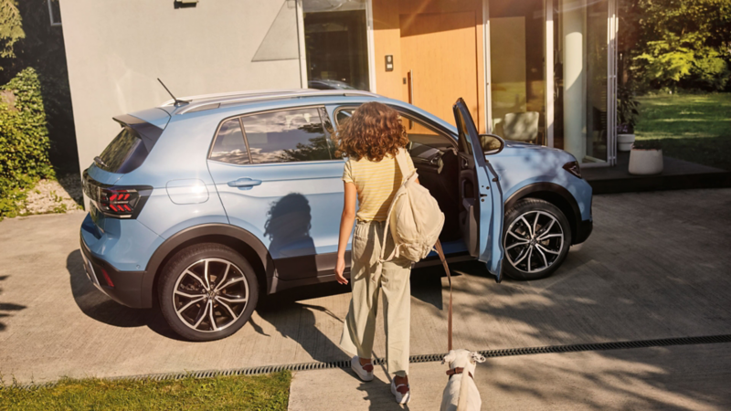 Une fille avec un chien se dirige vers un VW T-Cross bleu dans une allée. La porte passager est ouverte.