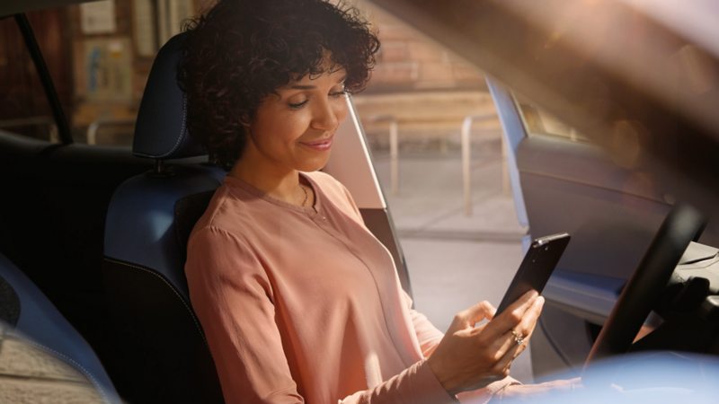 Eine lächelnde Frau sitzt auf dem Fahrersitz eines VW T-Cross und schaut auf ihr Smartphone.