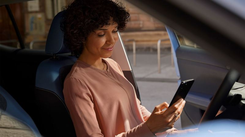 Une femme souriante regarde son téléphone portable sur le siège conducteur d'un VW T-Cross garé.