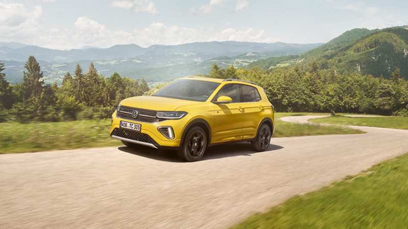 Ein gelber VW T-Cross fährt auf einer kurvigen Straße durch eine grüne Berglandschaft.