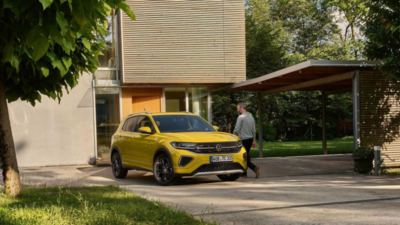 Ein gelber VW T-Cross steht in der Auffahrt eines modernen Wohnhauses mit großen Bäumen.