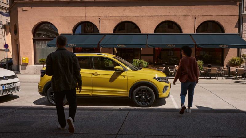 Mann und Frau nähern sich einem gelben VW T-Cross am Straßenrand.