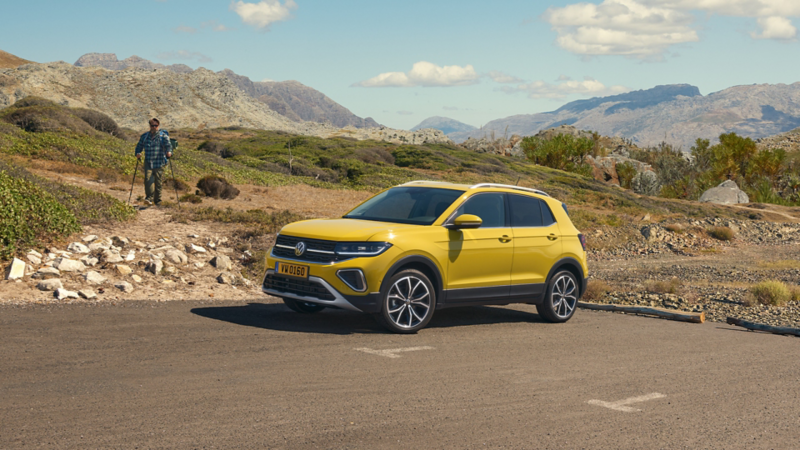 Une Volkswagen T-Cross jaune est garée sur le côté d'une route de campagne. À l'arrière-plan, un homme avec un sac à dos et des bâtons de randonnée marche sur un chemin de terre à travers un paysage accidenté de montagnes et de broussailles.