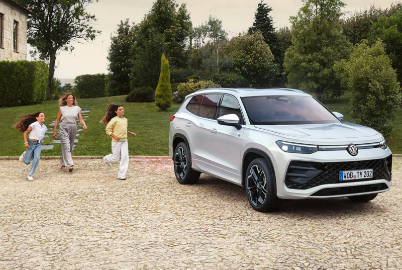 A smiling woman and two children run joyfully toward a parked white Volkswagen Tayron SUV on a cobblestone driveway, surrounded by lush greenery and a cozy house.