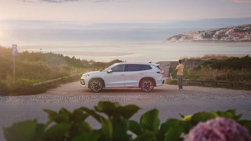 Ein Mann schaut über eine Düne auf das Meer im Sonnenuntergang, ein VW Tayron steht seitlich daneben.