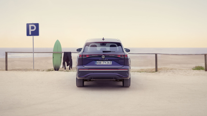 Ein grünes Surfbrett lehnt am Zaun eines sandigen Parkplatzes mit Meerblick neben einem VW Tayron.