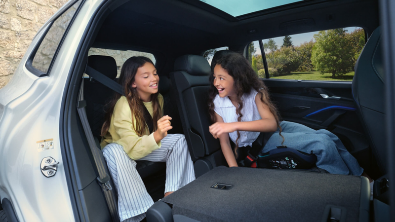 Two young girls sit in the back of a Volkswagen Tayron, smiling and chatting. One wears a yellow top, the other a white top. The scene exudes joy and friendship. Trees are seen through the car windows, indicating a rural setting.