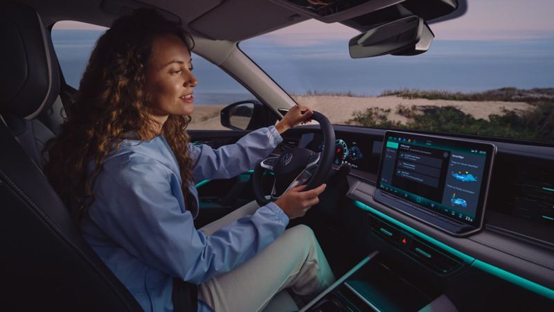 A woman smiling while driving a Volkswagen Tayron at dusk. The interior features illuminated dashboard and touchscreen displays. The mood is relaxing and serene.