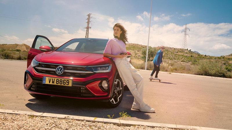 Une femme est appuyée contre le capot d'une Taiga accidentée. La portière côté passager est ouverte et derrière la voiture, un garçon fait du skateboard.