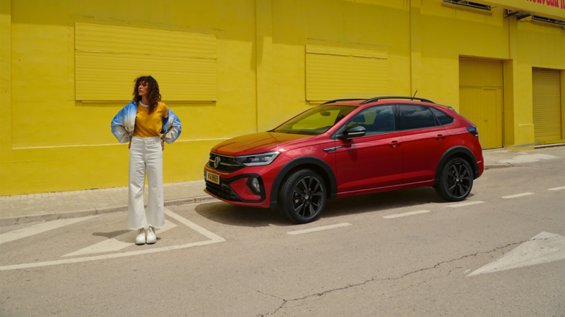 Une femme aux cheveux bouclés, vêtue d'une veste bicolore et d'un pantalon blanc, se tient à côté d'un SUV rouge garé devant un mur jaune.