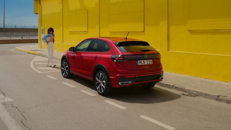 Röd VW Taigo parkerad längs vägen intill en gul vägg. Kvinna går framför bilen.