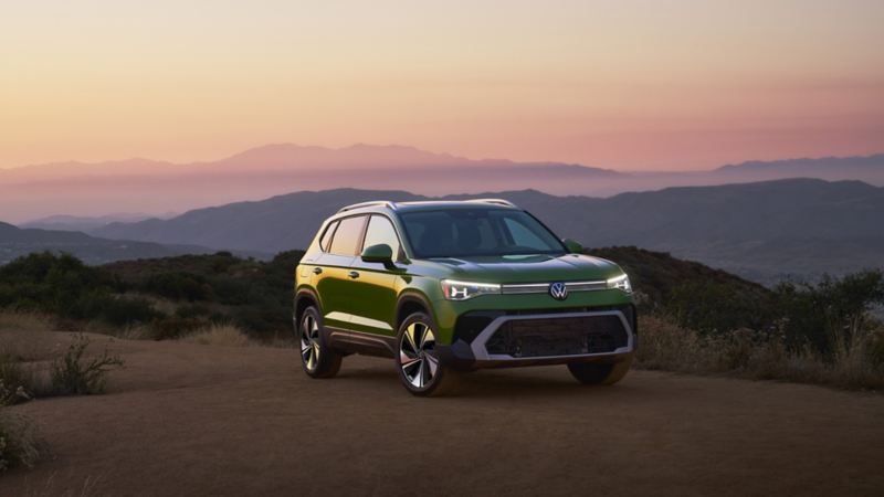 Un Taos 2025 vert stationné sur une piste en terre au crépuscule, devant un paysage de collines vallonnées et de montagnes au loin.