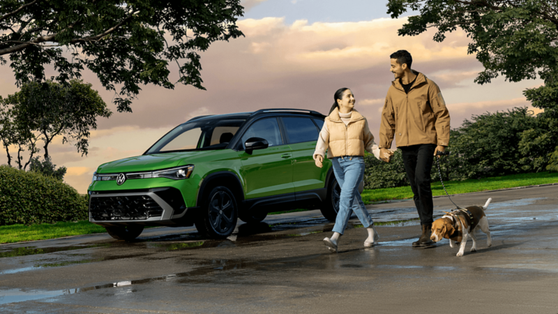 Un couple promenant son chien près d'un Volkswagen Taos 2025 vert garé dehors.