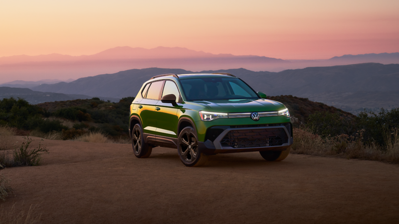 A green 2025 Taos parked on a dirt road during sunset against a backdrop of rolling hills and distant mountain ranges.