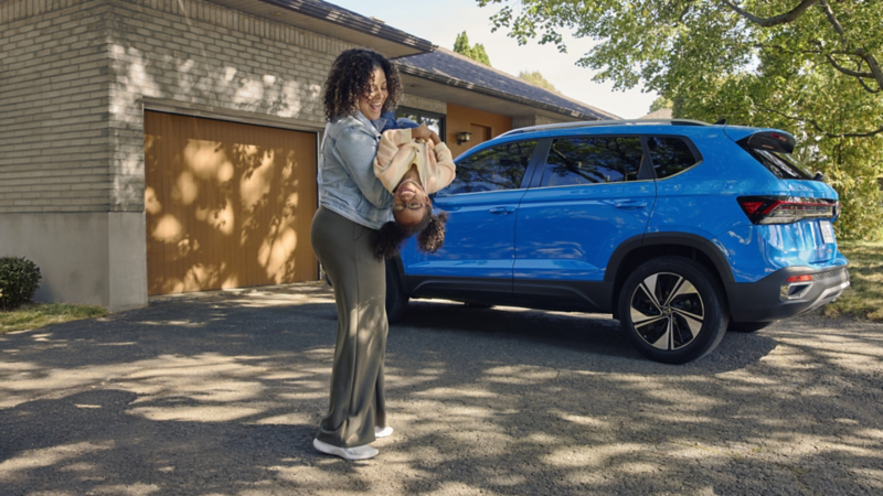 A woman carrying groceries walking toward a blue Volkswagen Taos parked in a residential driveway in front of a garage.