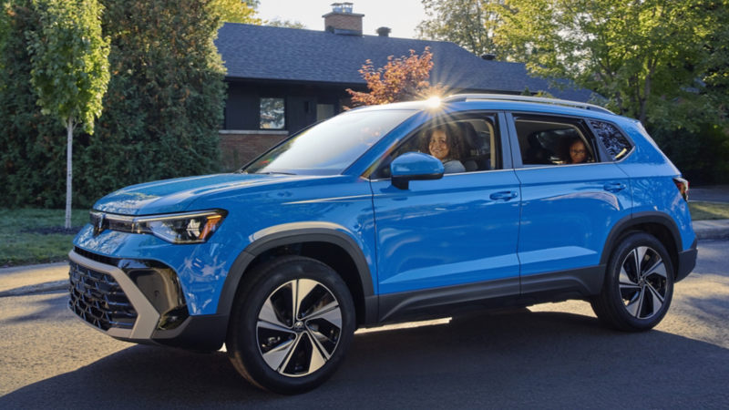 A blue Volkswagen Taos parked in a residential driveway in front of a house surrounded by trees.