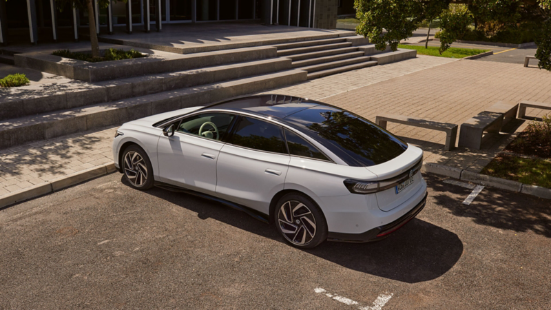 Une berline Volkswagen blanche dotée d'un toit panoramique noir, garée sur un parking devant un immeuble moderne doté d'un large escalier et d'espaces verts.