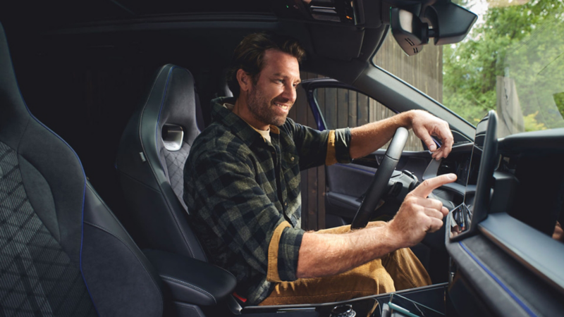 Un homme est assis au volant d'une voiture garée, souriant, et tape du doigt sur le système multimédia.