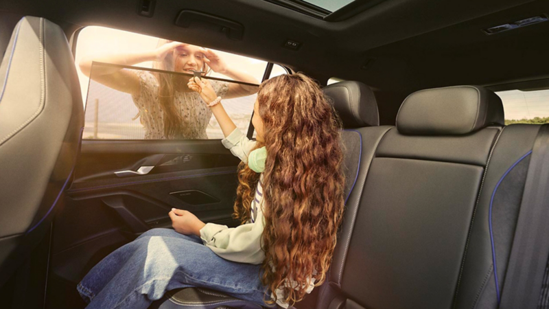Une fille est assise sur la banquette arrière et relève le pare-soleil de la fenêtre. De l'extérieur, une autre fille regarde à l'intérieur de la voiture.