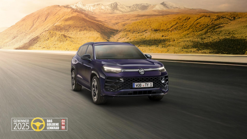 Ein lilafarbener VW Tayron fährt im Sonnenaufgang auf einer Straße durch die Landschaft. Ebenfalls abgebildet ist das Logo des Goldenen Lenkrads 2025.