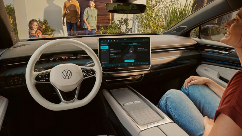 A woman sits in a modern Volkswagen car interior with a large touchscreen. Outside, a smiling family approaches, evoking a warm, welcoming atmosphere.