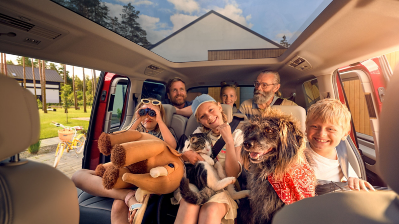 Intérieur d’un véhicule Volkswagen avec plusieurs personnes assises à l’arrière, dont des enfants, accompagnées d’un chien et d’un chat, avec des objets personnels, dans un cadre lumineux devant une maison.