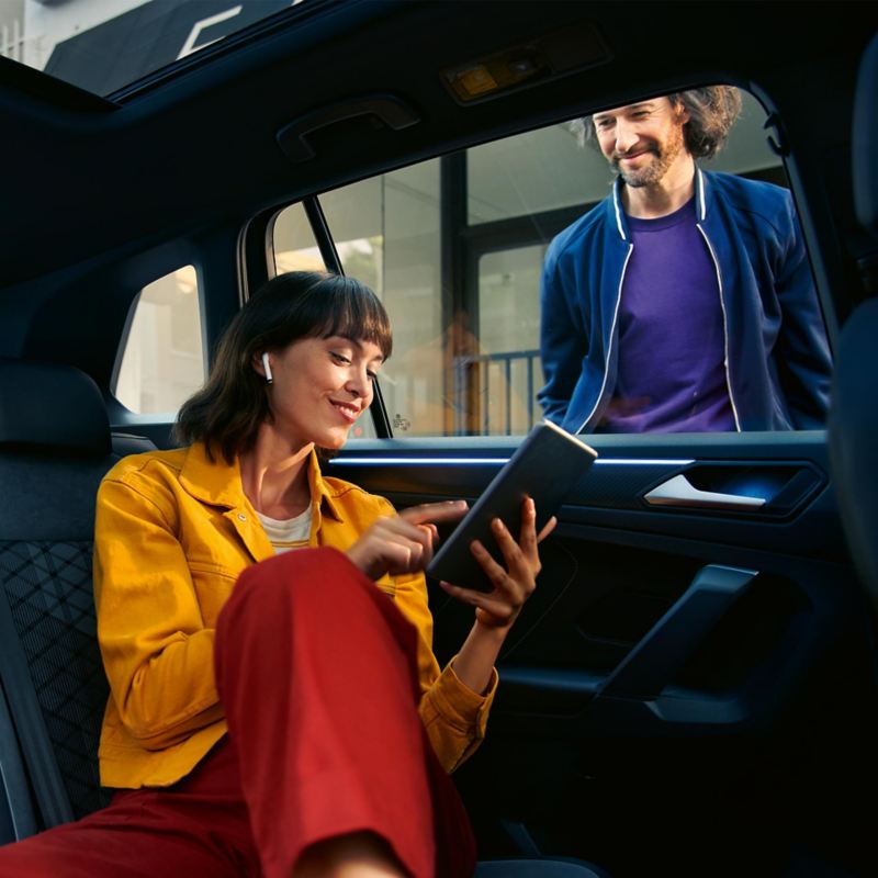 Μια γυναίκα κάθεται στο πίσω ενιαίο κάθισμα ενός οχήματος VW Tiguan και χρησιμοποιεί το We Connect από το tablet της