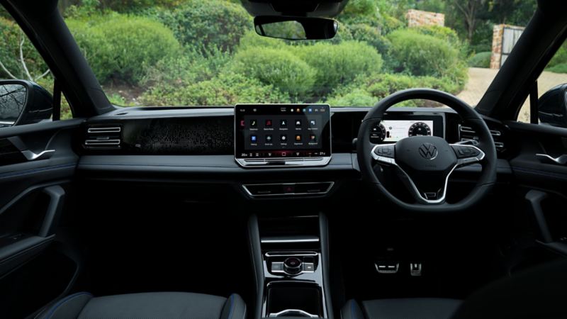 A young man on the driver side of the dark green VW Tiguan operates the optional Park Assist Plus from his smartphone.