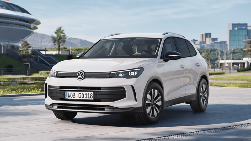 Biały Volkswagen Tiguan stoi zaparkowany przed nowoczesnym stadionem, w tle góry i miasto.
