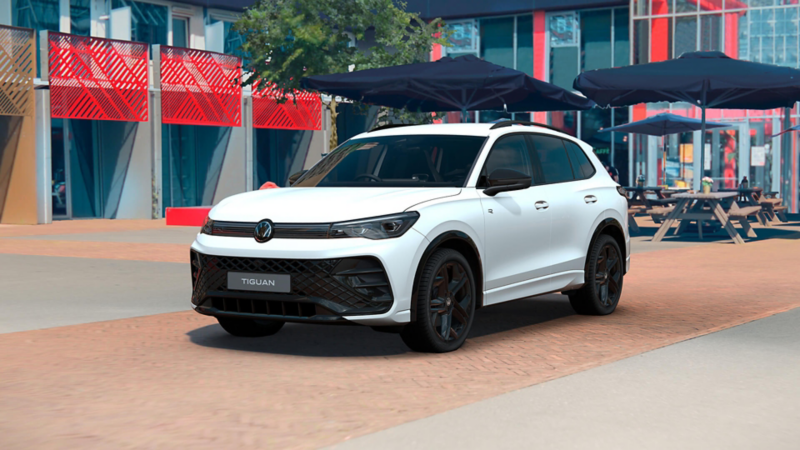 A white Volkswagen Tiguan Black Edition is parked on the roadside in front of a restaurant with outside eating benches and umbrellas 
