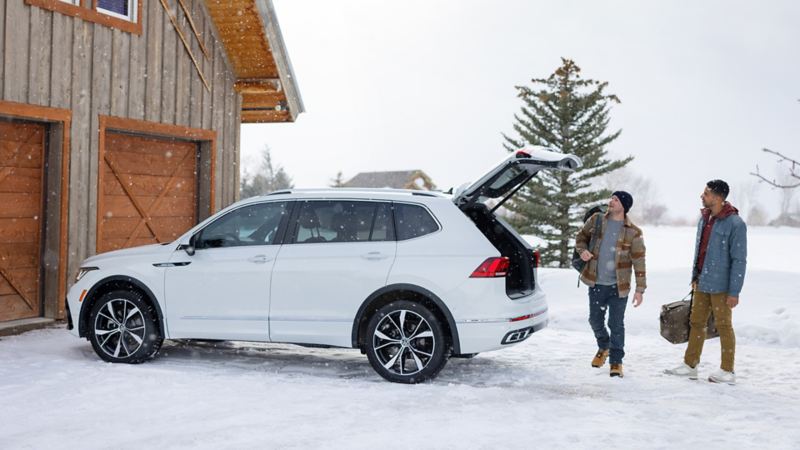 2 men taking bags from the open trunk of the white 2024 Volkswagen Tiguan parked near a wooden house