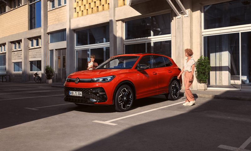 Na zdjęciu widać czerwonego Tiguana marki Volkswagen zaparkowanego na zewnętrznym parkingu przy nowoczesnym budynku, a obok niego stoi kobieta w jasnym stroju.