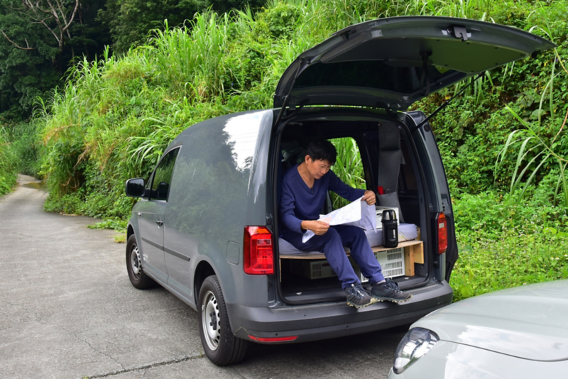 鏡色灰的Caddy Van停在路旁，車尾門掀起後，車主坐在自行加裝的床墊上看手上的文件