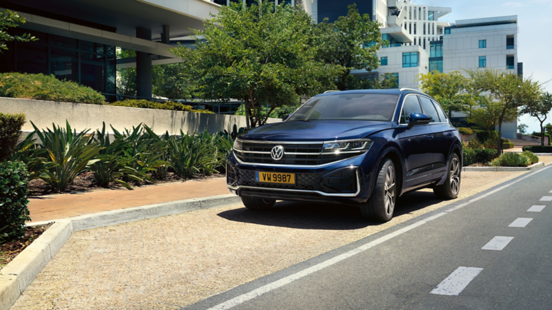 Ce plan en plongée montre un Volkswagen Touareg R-Line bleu foncé, garé dans une rue. L'avant de la voiture est visible. Des bâtiments modernes se trouvent en arrière-plan, ainsi que des arbres. 