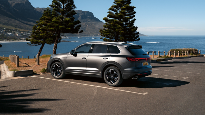 Un SUV Volkswagen Touareg gris foncé est garé sur un parking en bord de mer. La voiture est vue de trois quarts arrière, avec les vagues de l'océan et des arbres en arrière-plan. La carrosserie est élégante et les jantes noires sont mises en valeur.