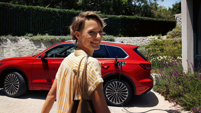 Une femme souriante, portant une chemise rayée et un sac marron, regarde par-dessus son épaule en marchant. Derrière elle se trouve une Volkswagen Touareg rouge, branchée à une borne de recharge.