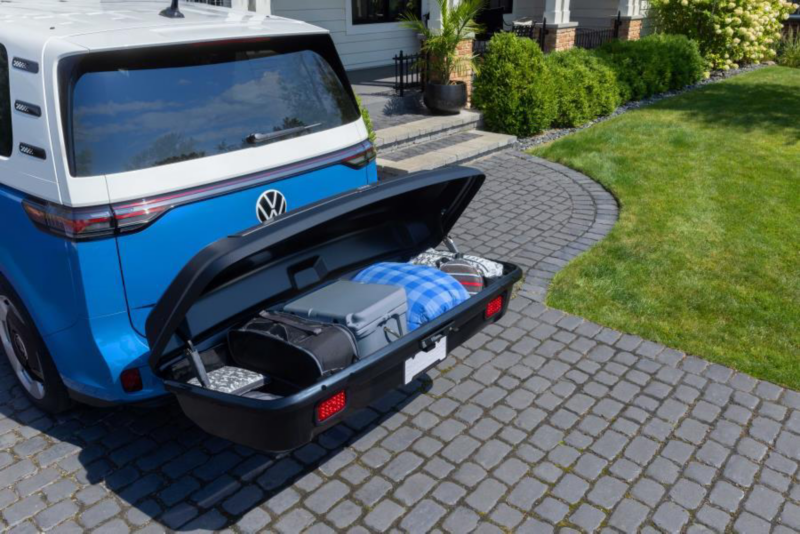 Coffre de rangement arrière ouvert monté sur une Volkswagen bleue, rempli de valises et sacs de voyage, stationnée sur une allée pavée. 