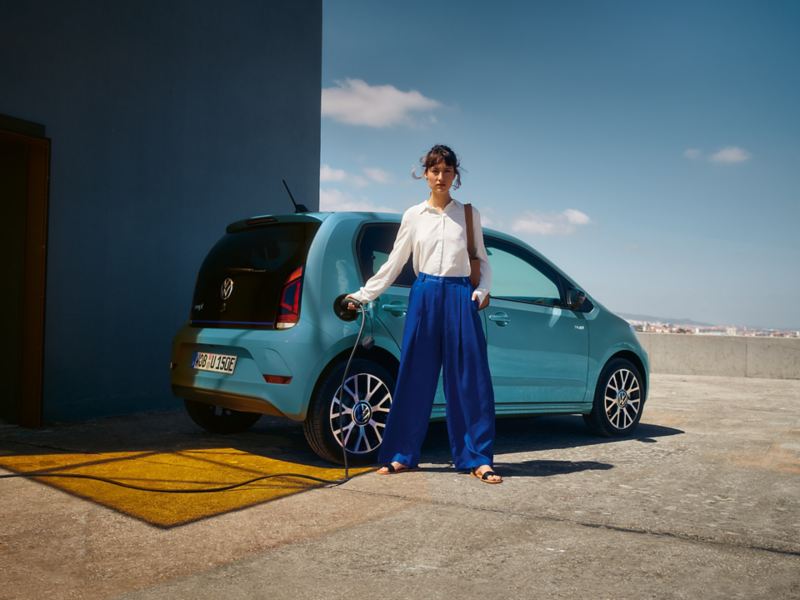 VW e-up! Auto seisoo pysäköintitalon katolla ja latautuu latauskaapelilla, jota nainen on liittämässä pistorasiaan.