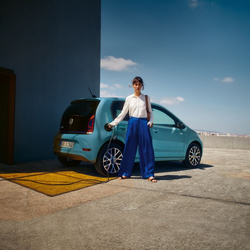 VW e-up! Se encuentra en el techo de un aparcamiento, se carga con un cable eléctrico que una mujer enchufa a la toma de corriente.