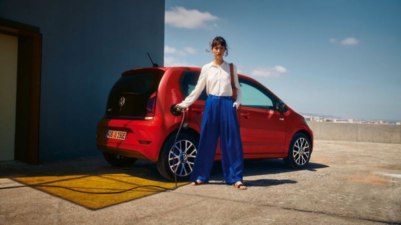 Roter VW e-up! steht auf Parkhausdach und wird mit Stromkabel geladen, welches von einer Frau in die Steckdose gesteckt wird.