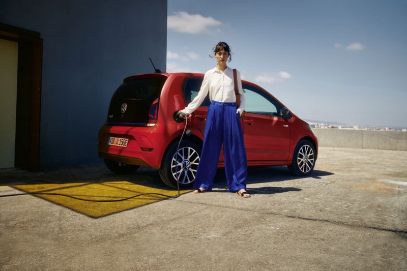 Una ragazza inserisce il cavo di ricarica nella Volkswagen Nuova e-up! vista 3/4 posteriormente e parcheggiata in uno spiazzo.