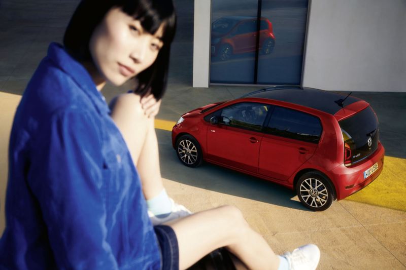 Roter VW e-up! steht vor Haustür, Blick von oben auf Dach, Seite und Heck. Dunkelhaarige Frau sitzt im Vordergrund auf Mauer.