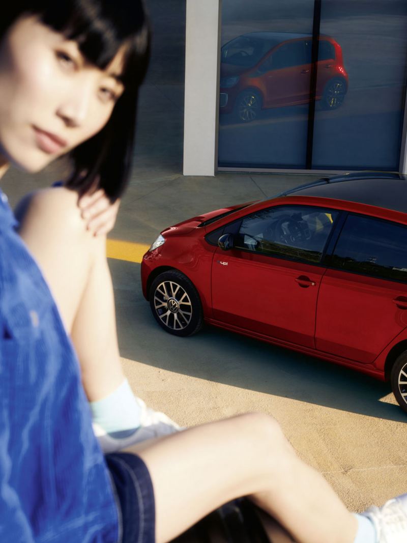 Roter VW e-up! steht vor Haustür, Blick von oben auf Dach, Seite und Heck. Dunkelhaarige Frau sitzt im Vordergrund auf Mauer.