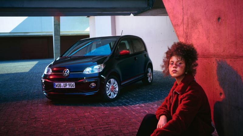 Schwarzer VW up! "beats" mit roten Spiegelkappen in Parkhaus, Blick auf Front, Frau in roter Kleidung sitzt davor.