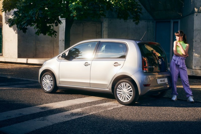 Una ragazza in piedi dietro a Volkswagen eco-up!, vista 3/4 posteriormente e parcheggiata a bordo di una strada cittadina.