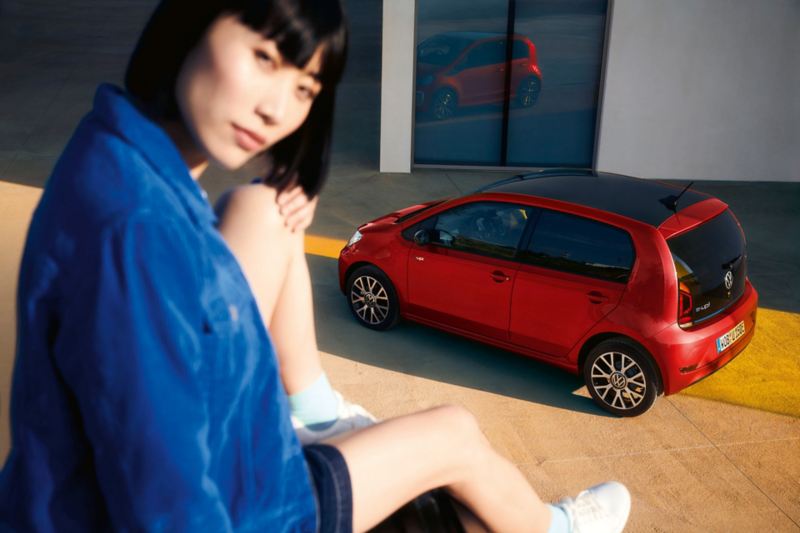 Roter VW e-up! Edition steht vor Haustür, Blick von oben auf Dach, Seite und Heck. Dunkelhaarige Frau sitzt im Vordergrund auf Mauer.