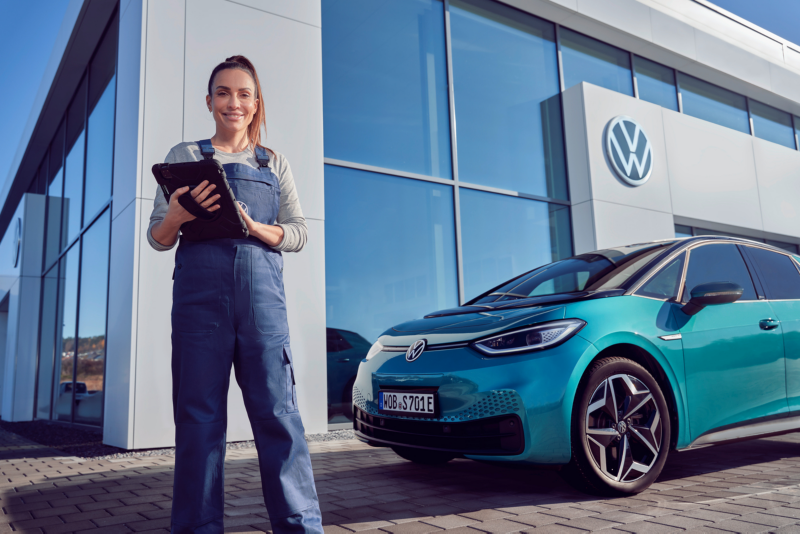 VW employee standing outside a dealership holding a clipboard and next to a blue ID.3